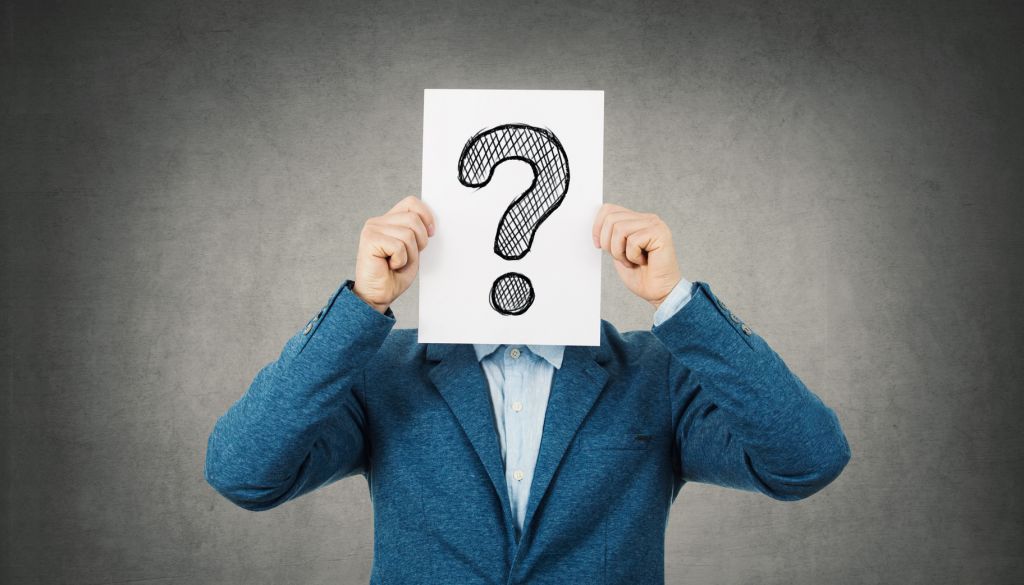 Businessman covering his face using a white paper with drawn question mark, like a mask, for hiding his identity. Isolated gray wall background.