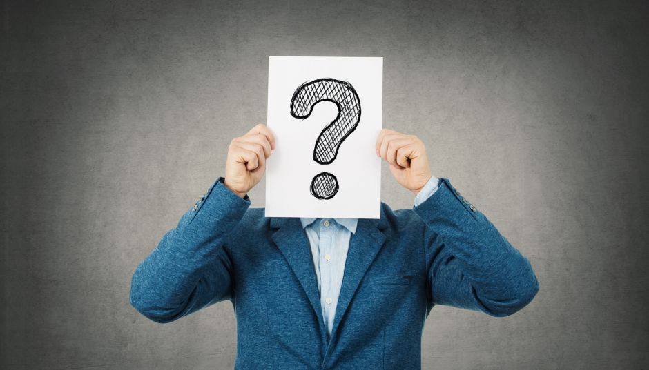 Businessman covering his face using a white paper with drawn question mark, like a mask, for hiding his identity. Isolated gray wall background.